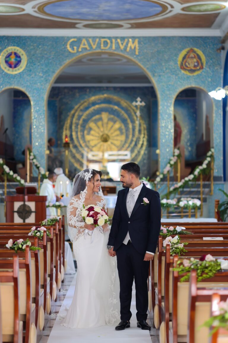 A couple at the head of the aisle in a church with a gold and blue mosaic apse, the pews dressed with pink and white flowers, Clorinda Bellone Elite Weddings