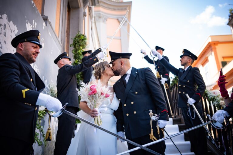 A couple kiss beneath a sword arch formed by a uniformed guard of honour at the foot of the church steps, Clorinda Bellone Elite Weddings