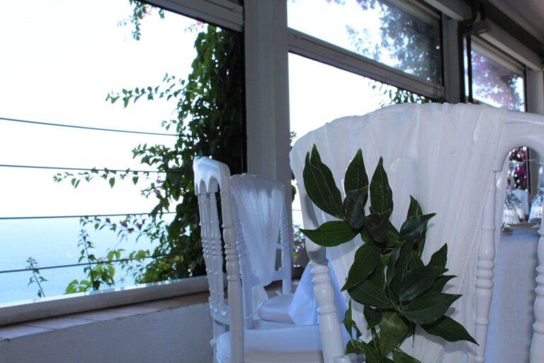 A white chair dressed with a sprig of bay leaves beside windows over the sea, Clorinda Bellone Elite Weddings