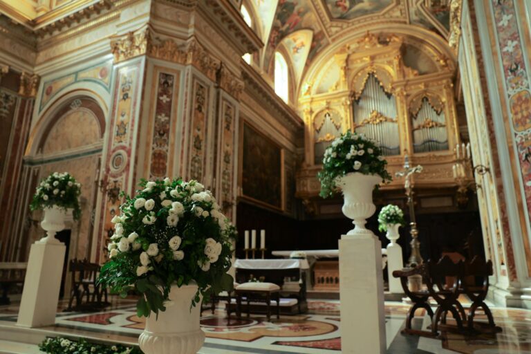 Urns of white roses on white pedestals before the altar and gilded organ of a Baroque church, Clorinda Bellone Elite Weddings