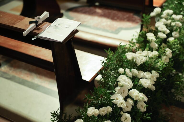 White roses and greenery along the end of a pew, with an order of service and fan left on the bench, Clorinda Bellone Elite Weddings
