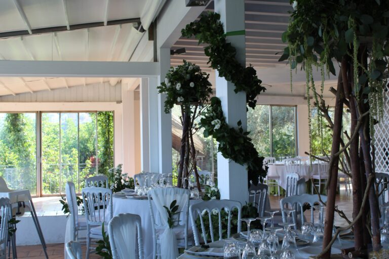 A glazed terrace dining room before guests arrive: white chairs, climbing greenery and tree-trunk stands of white flowers, Clorinda Bellone Elite Weddings