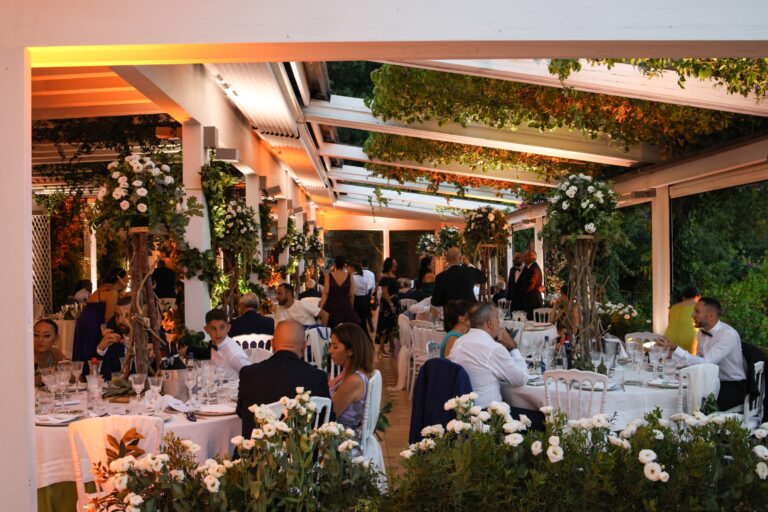 An evening reception under a lit pergola, with borders of white roses and floral columns between the tables, Clorinda Bellone Elite Weddings