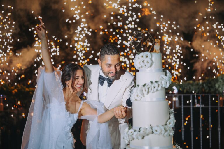 Cutting a three-tier white cake against a curtain of fairy lights, Clorinda Bellone Elite Weddings