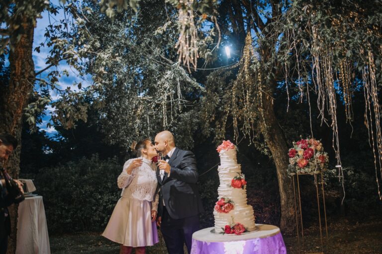 A toast beside a tiered cake beneath trees hung with lights, with a stand of red and pink roses, Clorinda Bellone Elite Weddings