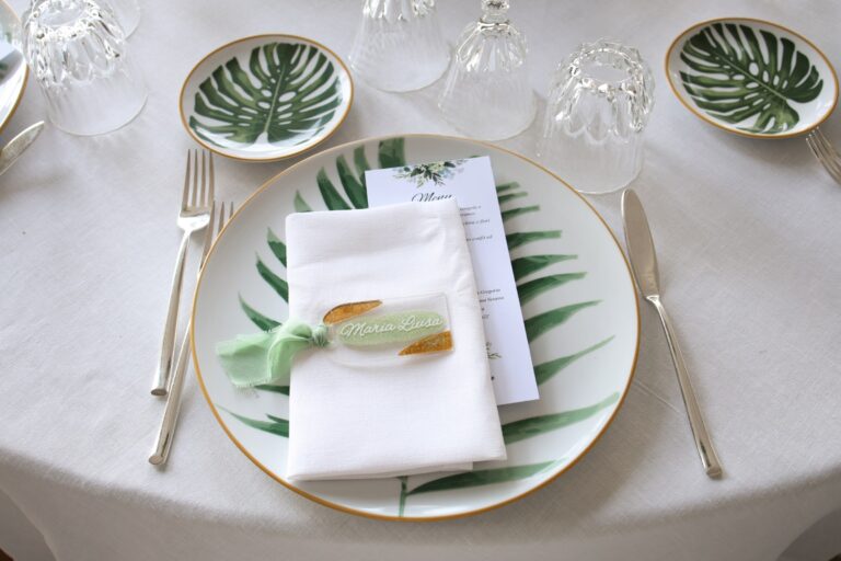 A place setting with monstera-leaf china, a linen napkin, a printed menu and a name tag tied with green ribbon, Clorinda Bellone Elite Weddings