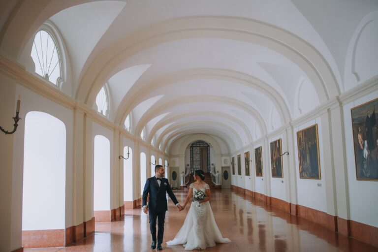 A couple walk hand in hand along a long vaulted gallery hung with paintings, Clorinda Bellone Elite Weddings
