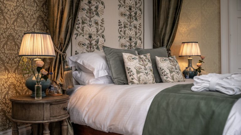 A bedroom with patterned wallpaper, lamps on either side of the bed and embroidered cushions, Cluny Castle