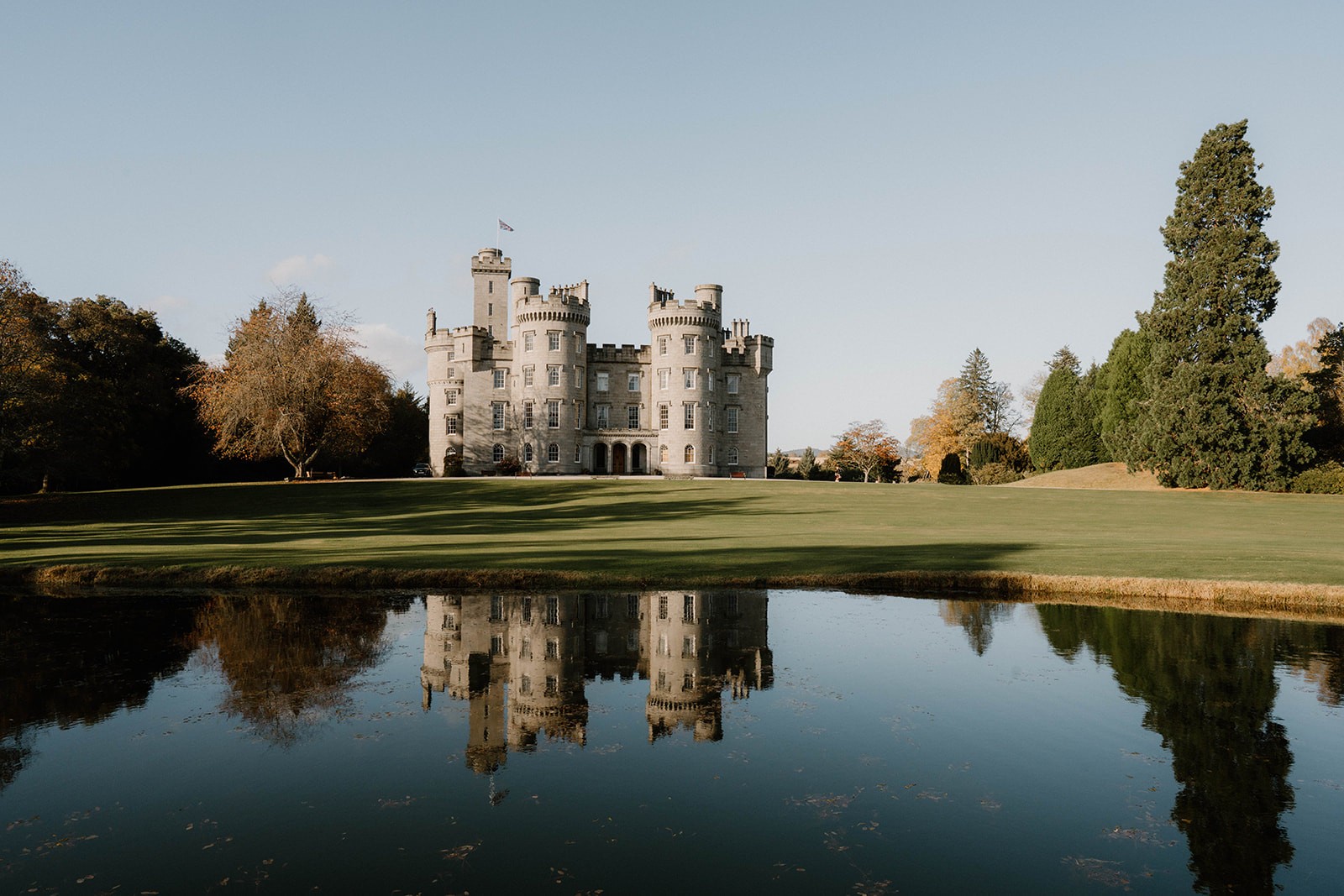 The castle reflected in still water from the far side of the pond, with autumn colour in the trees, Cluny Castle