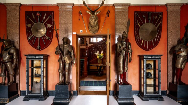 A hall of suits of armour, weapon displays and a stag's head around a doorway to the staircase, Cluny Castle