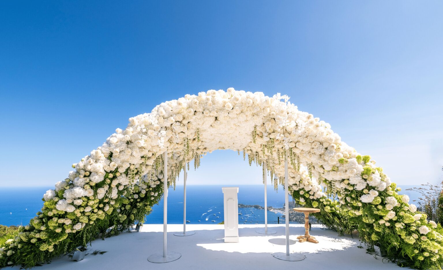 A floral ceremony arch on a white platform above the sea