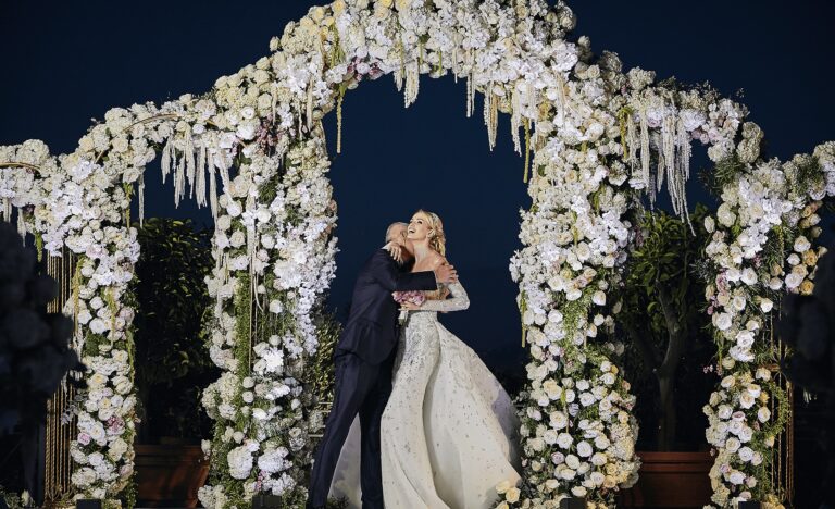 Couple embracing beneath a white floral arch at night, Cocoon Events
