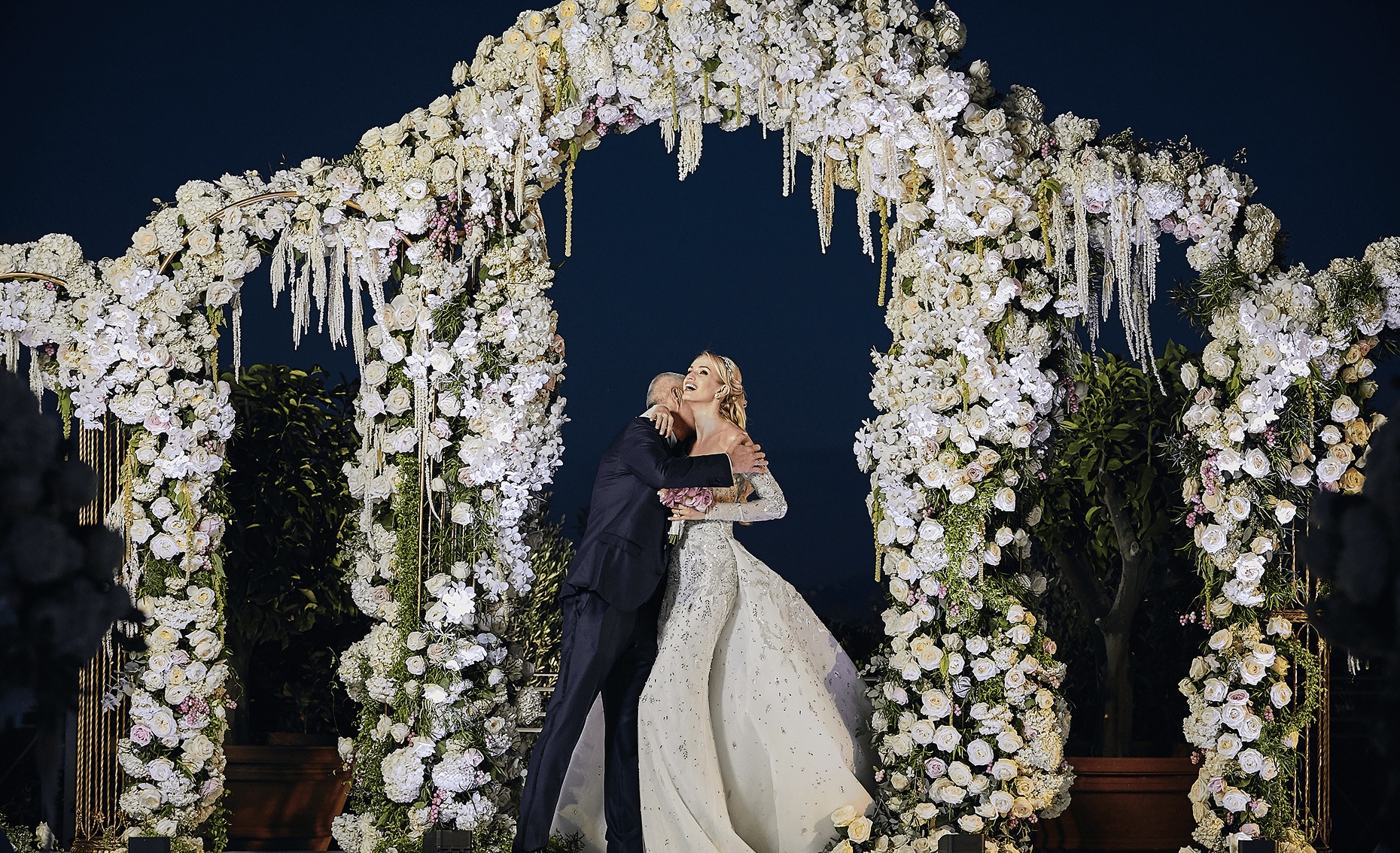 Couple embracing beneath a white floral arch at night, Cocoon Events