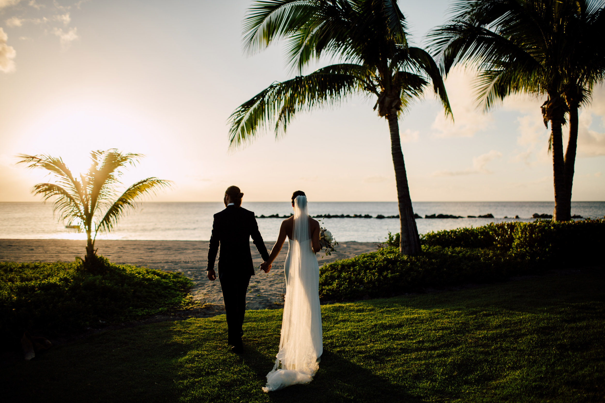 The couple walking towards the sea at sunset under palms, from their Caribbean destination wedding, Cottonwood Studios