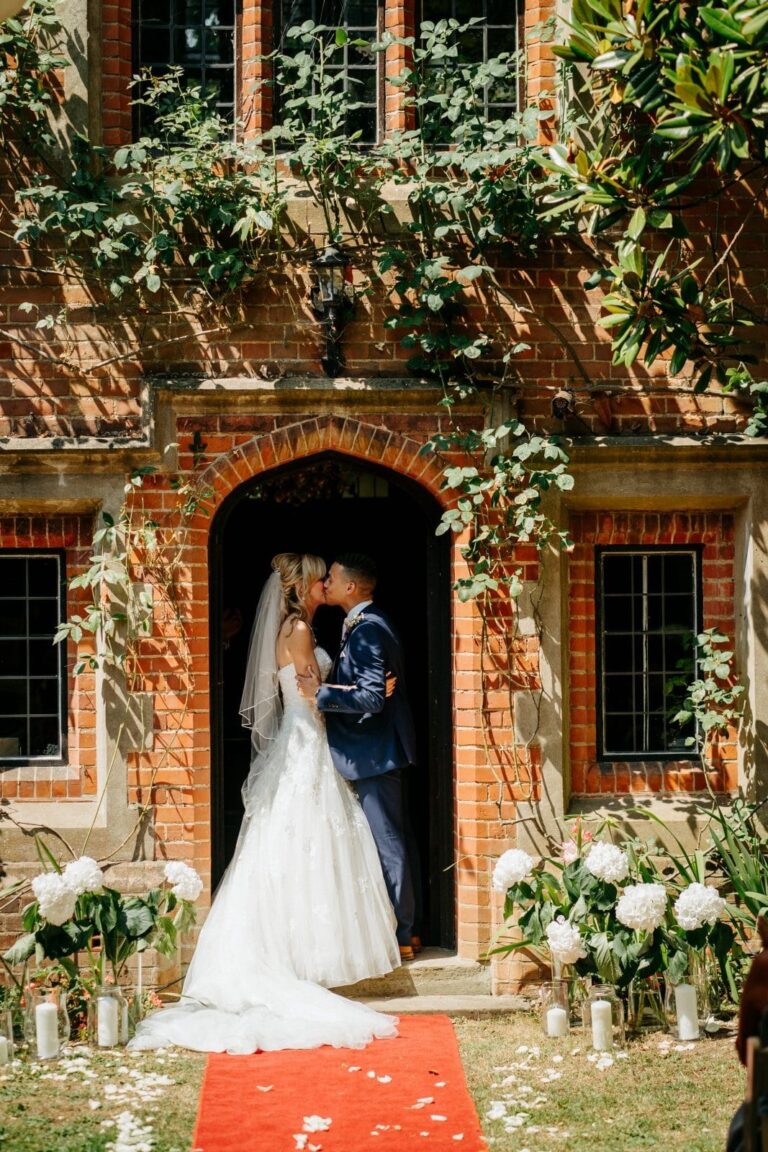The couple kissing in the Tudor arched doorway, red carpet and floral urns, Creeksea Place