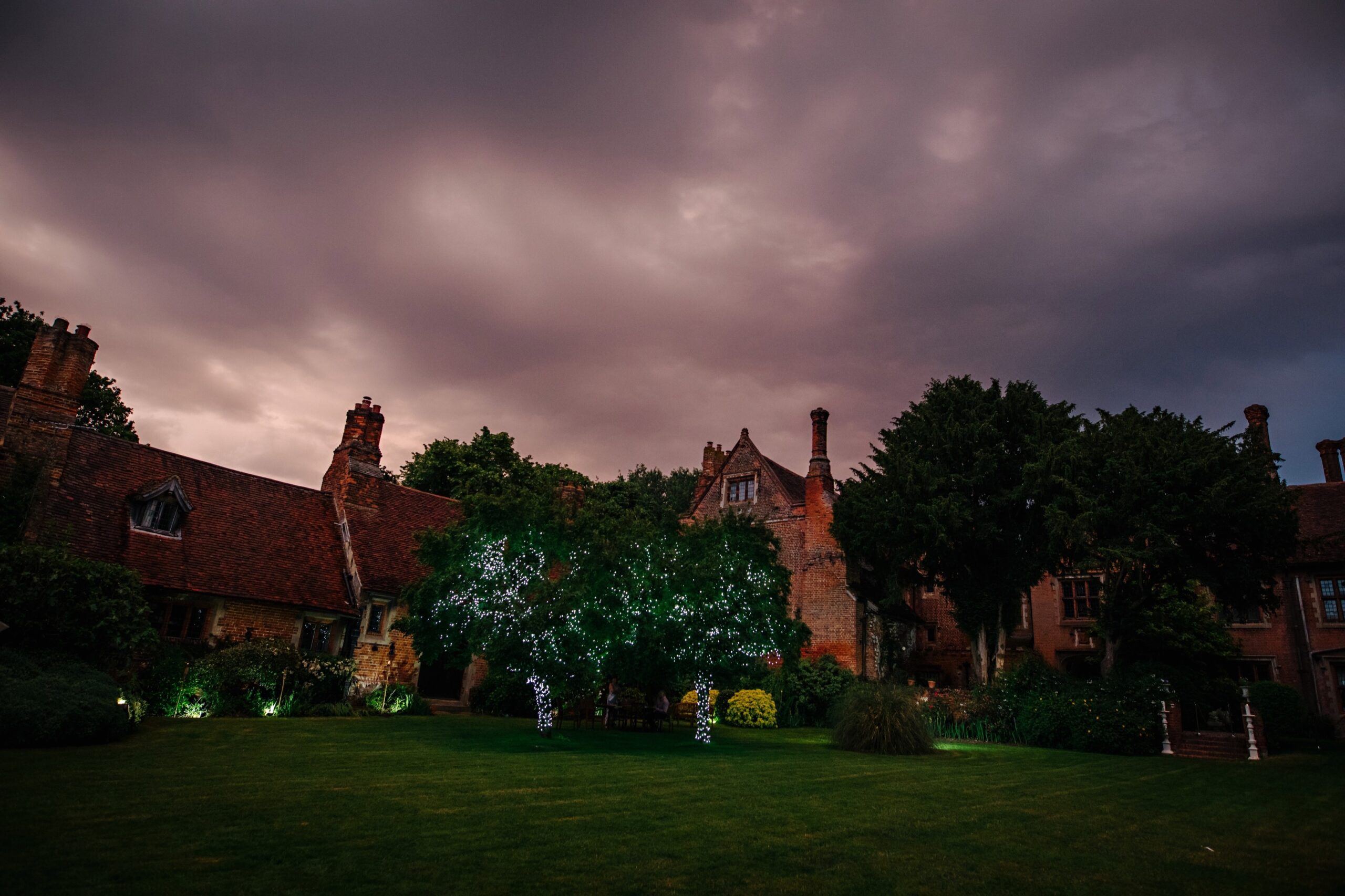 The manor at dusk under a violet sky, chimneystacks and fairy-lit trees over the lawn, Creeksea Place