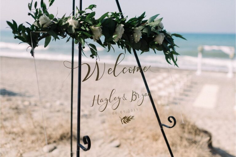 A welcome sign with an olive garland on the sand in front of ceremony chairs, Crete for Love