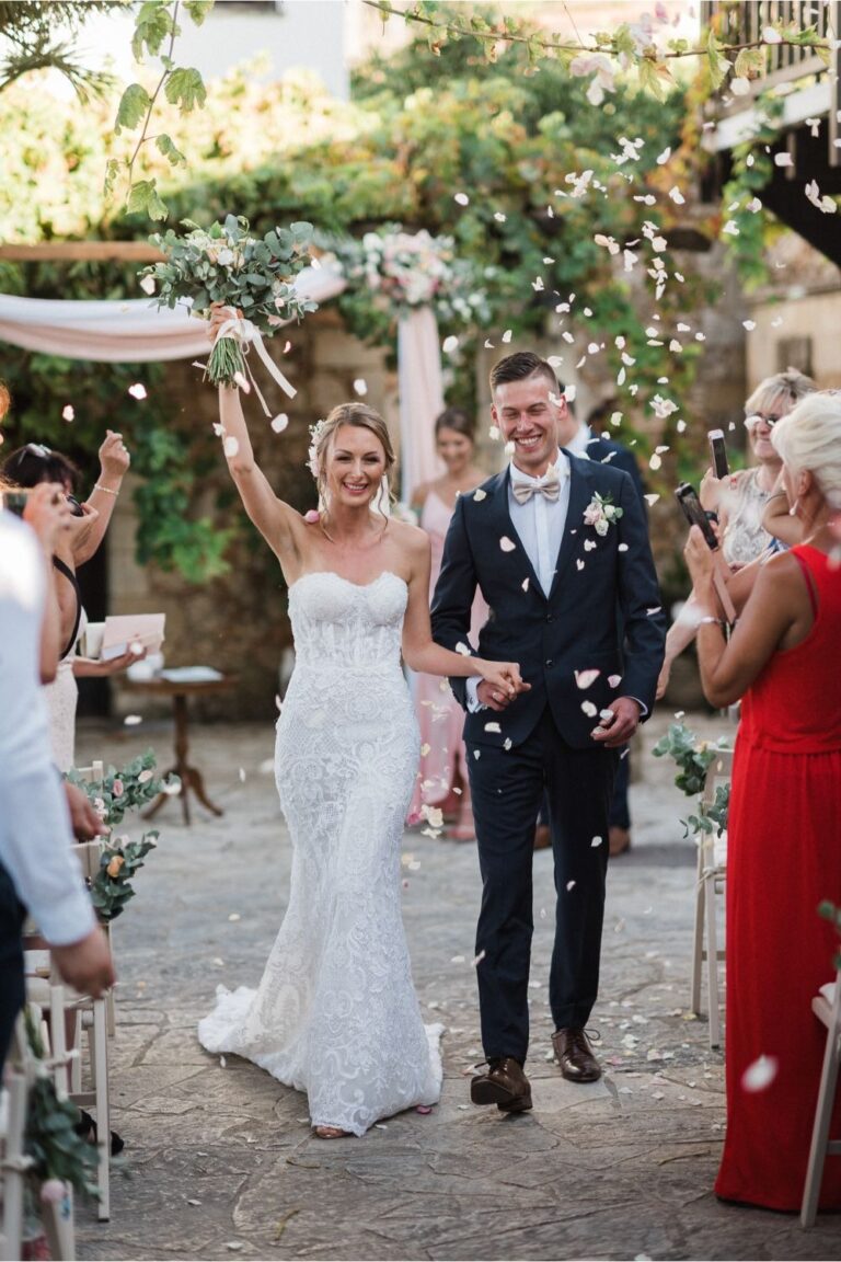 A couple walk down a garden aisle as guests throw petals, Crete for Love