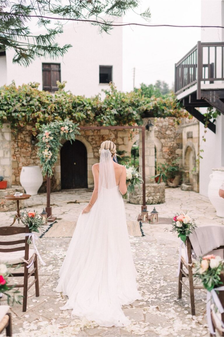 A bride in a long veil walks towards a courtyard ceremony set up under a vine, Crete for Love