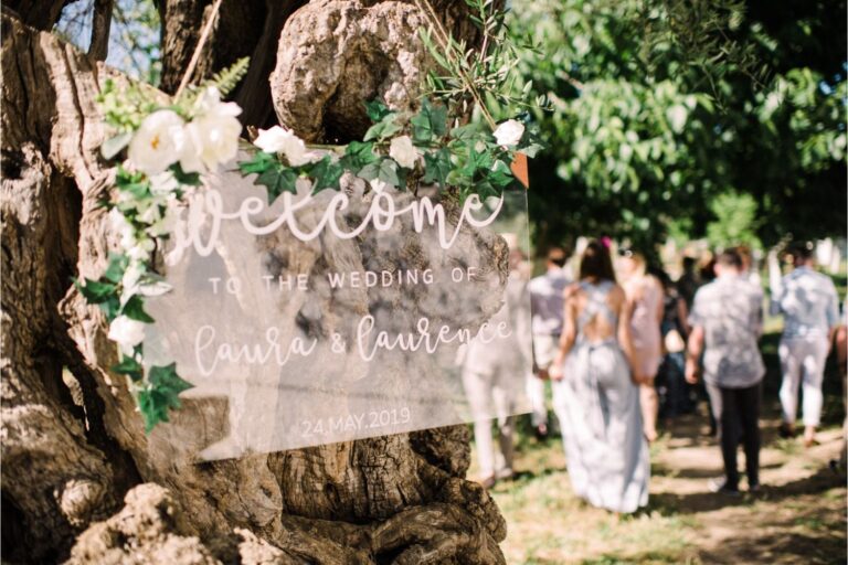 A transparent welcome sign decorated with white flowers hangs on an olive tree, Crete for Love
