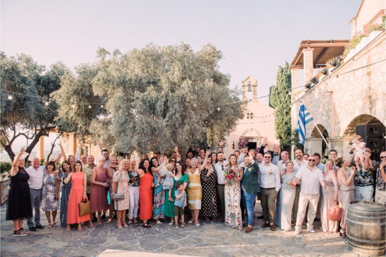 Wedding guests in bright outfits gather for a group photograph in a stone courtyard, Crete for Love