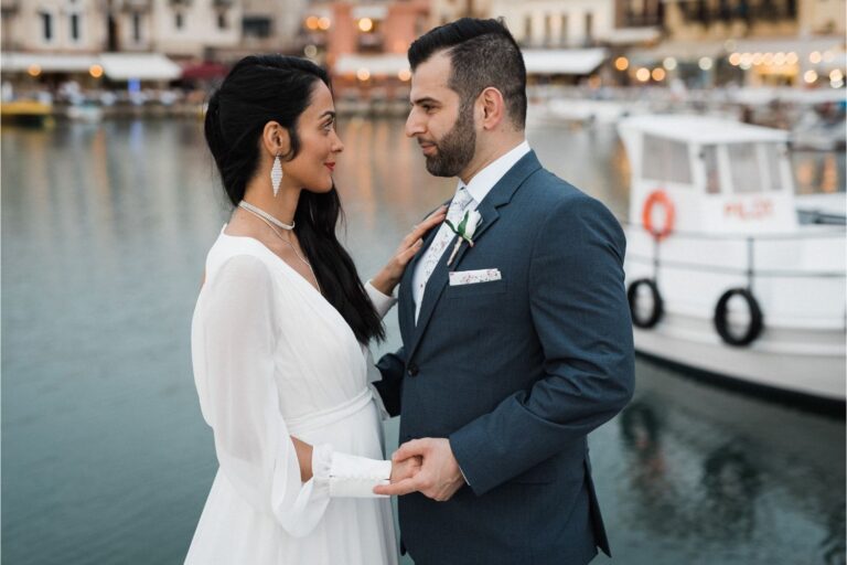 A couple hold hands beside a harbour with moored boats, Crete for Love