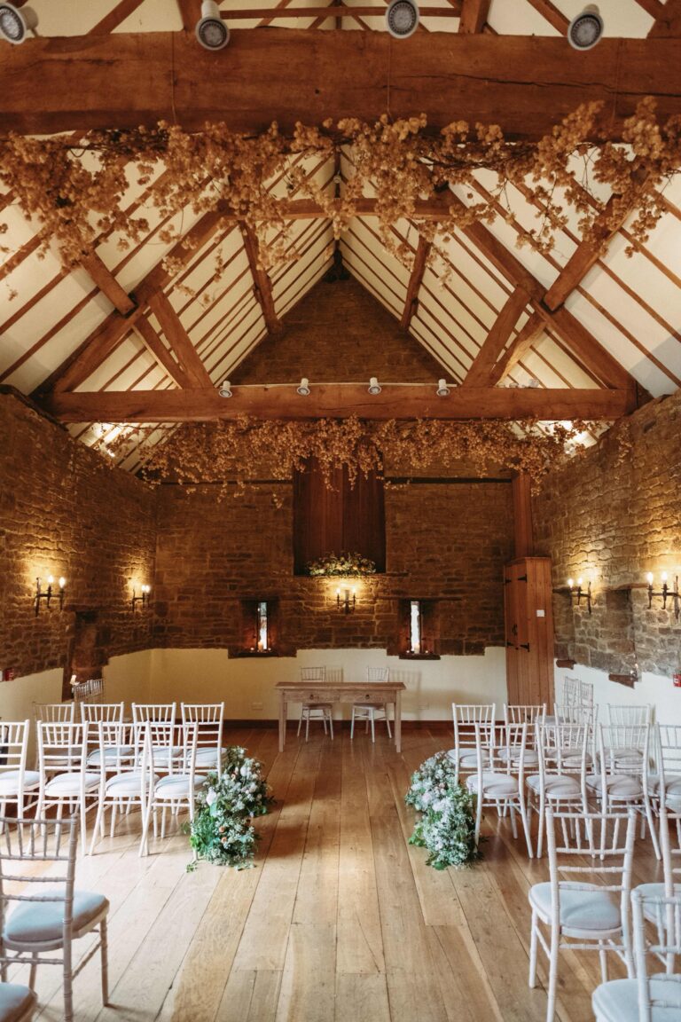 The vaulted stone barn set for a ceremony, with oak roof trusses, candles and white chairs either side of the aisle, Crockwell Farm