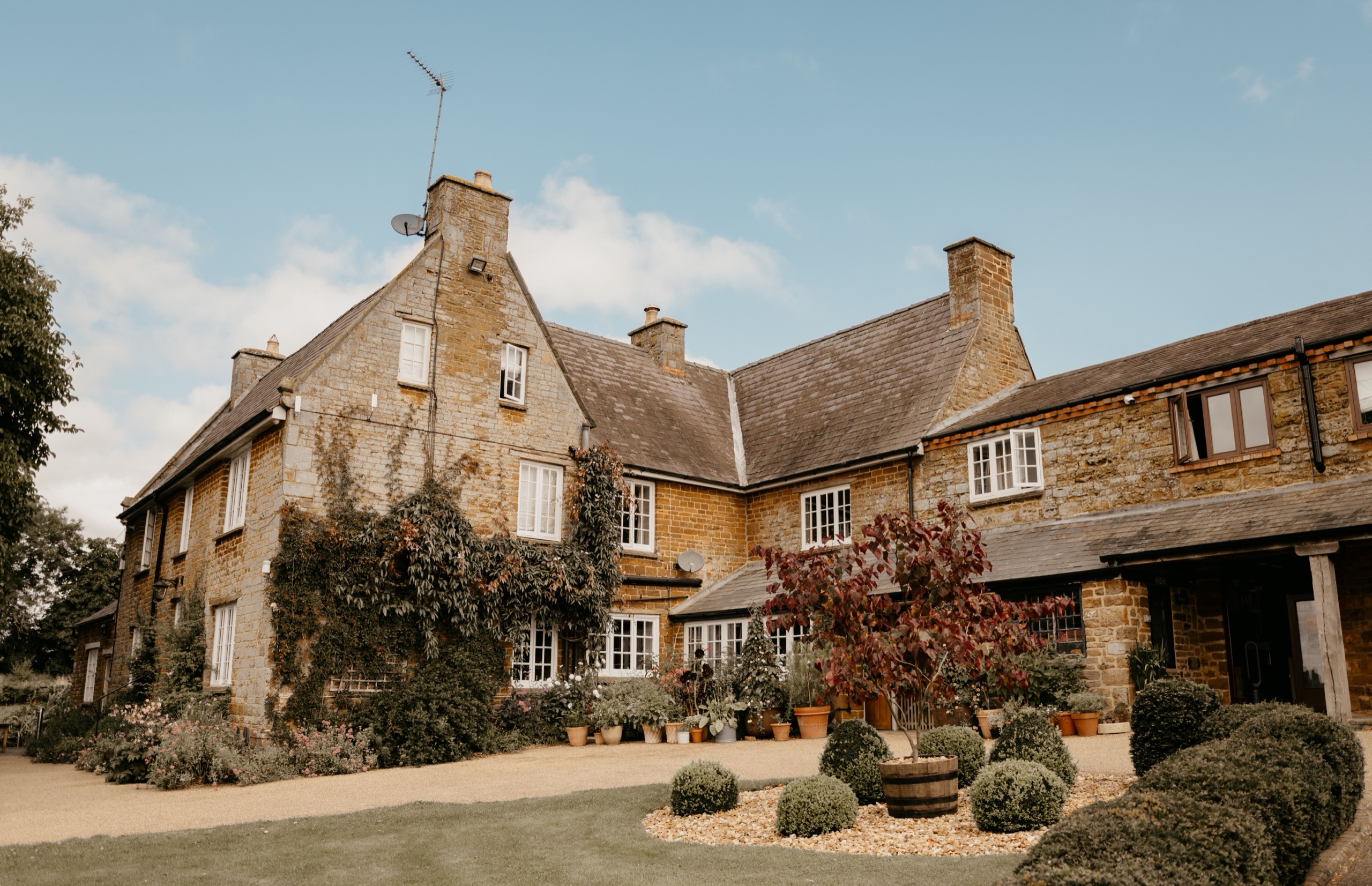 The ironstone Farmhouse and its gravel forecourt, with climbers on the walls and clipped box in the front garden, Crockwell Farm