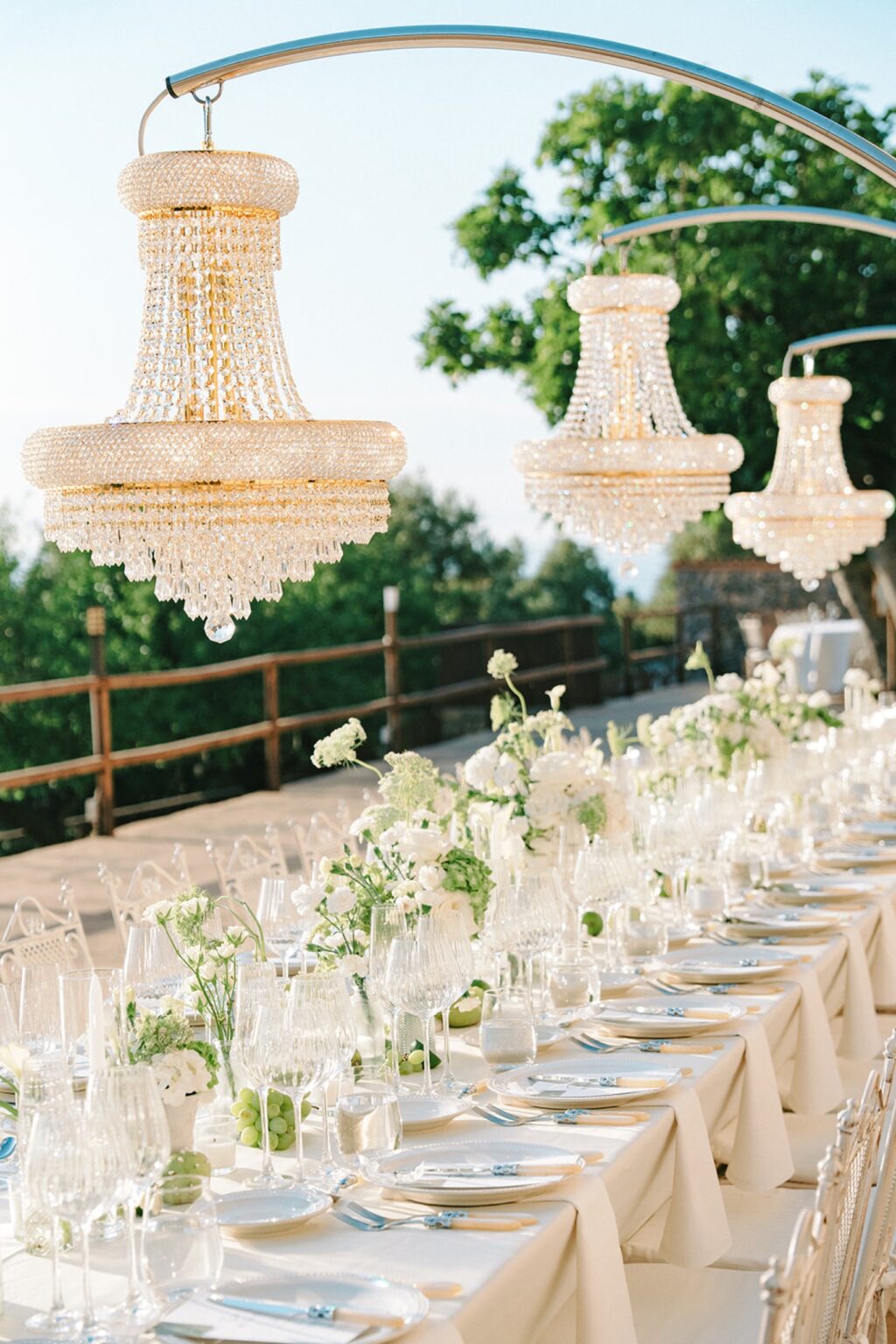 A long table dressed in white under hanging crystal chandeliers on a terrace