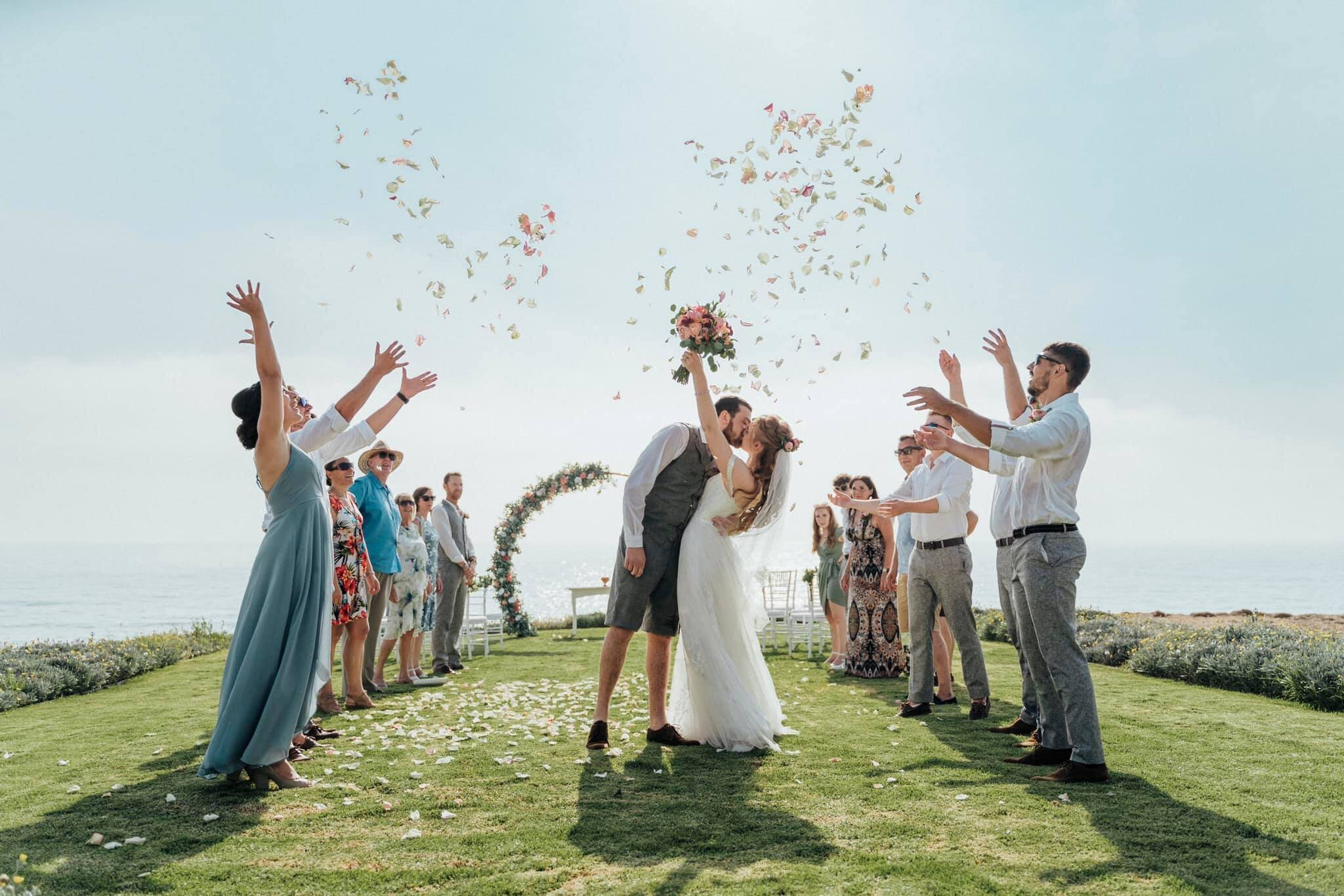 Guests throwing petals as the couple kiss beside a floral ring on a clifftop lawn, Cyprus Wedding Touches