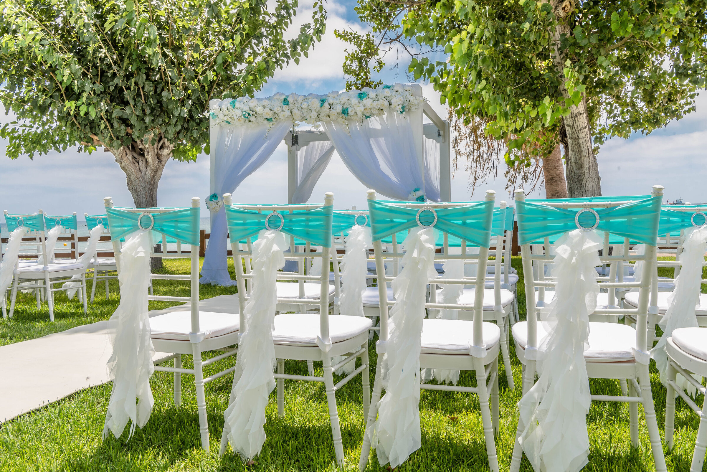 Wedding canopy and aisle with turquoise-sashed chairs beneath shade trees, Cyprus Wedding Touches