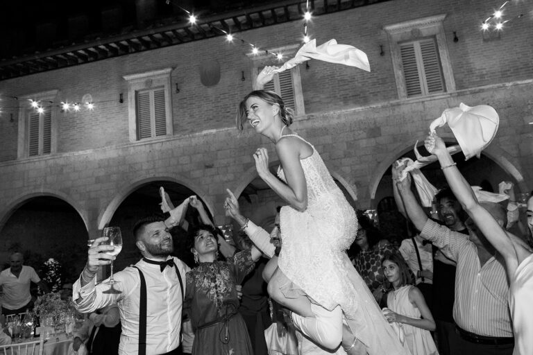 The bride lifted onto guests' shoulders, napkins in the air, in a lit courtyard in Chianti, black and white, Daniele Vertelli Photography