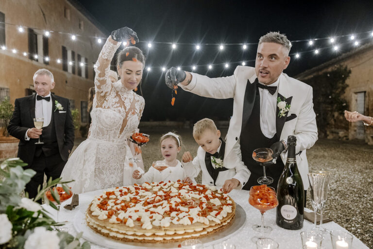 Bride and groom scattering strawberries over a millefoglie wedding cake under string lights, children helping, Villa di Catignano near Siena, Daniele Vertelli Photography
