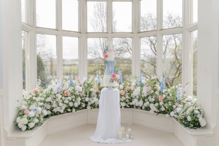 A tall wedding cake with sugar flowers in a bay window ringed with flowers, Danielle's Cakes Pershore