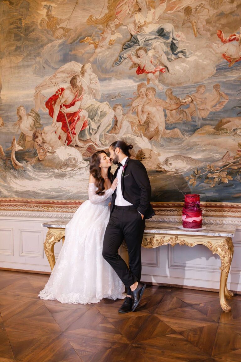 Couple beside a red petal-layered wedding cake in a tapestry-hung salon, Daria Events and Weddings