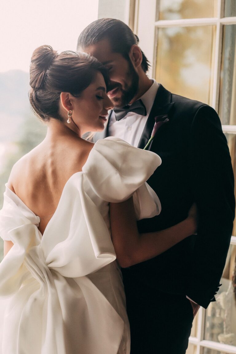 Couple embracing by a tall window, bride in a bow-backed gown, Daria Events and Weddings