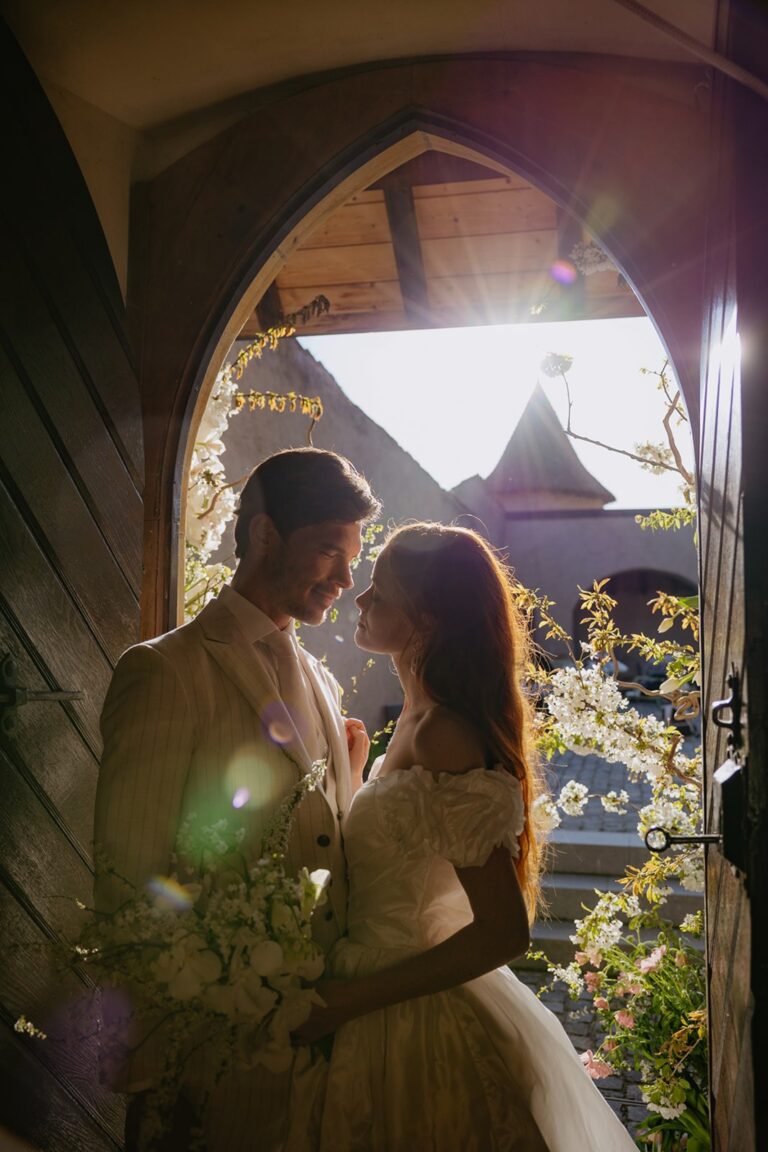 Couple framed in an arched wooden doorway with a chapel behind, Daria Events and Weddings