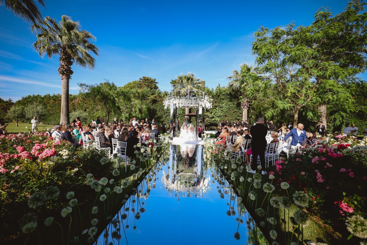 Ceremony aisle laid over mirrored water between flower beds and palms in a garden (venue not named by the planner), Dario Varsalona