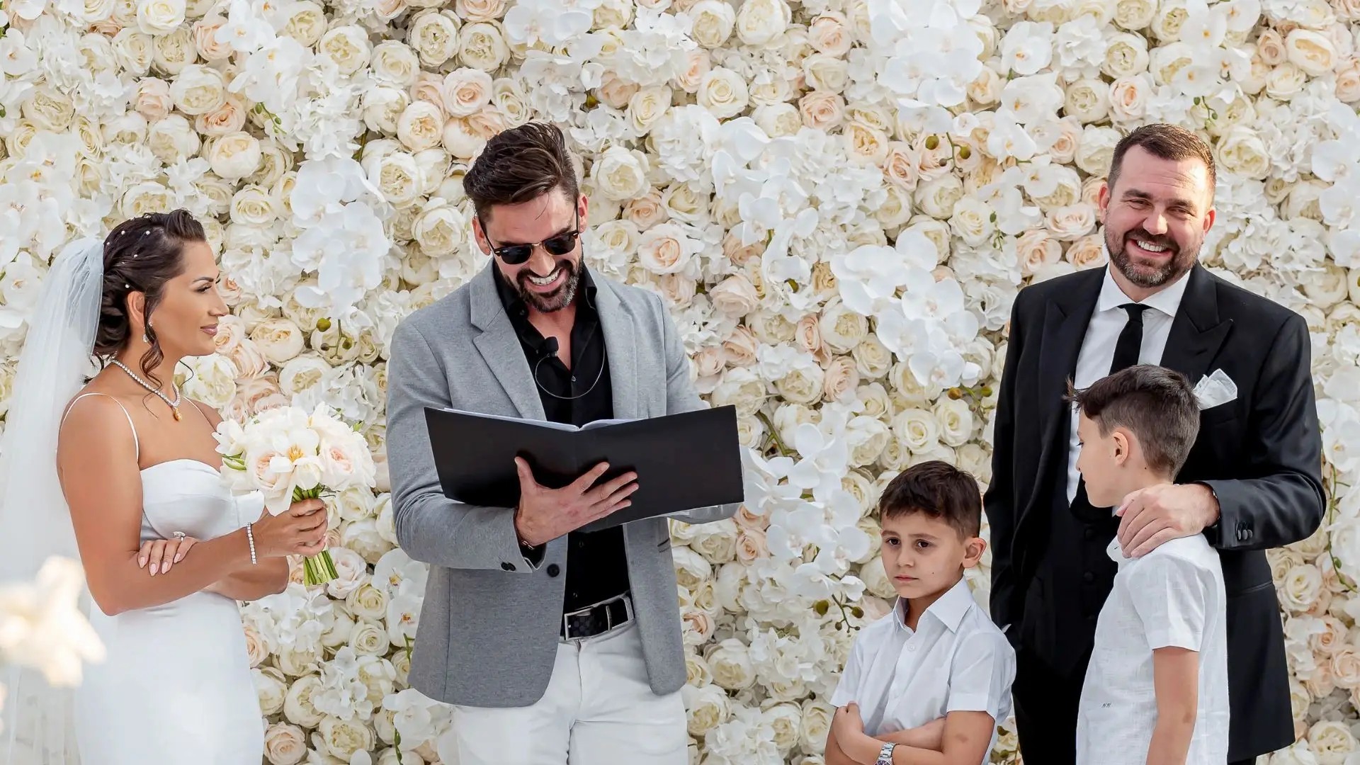 Celebrant with a bride and a groom with two children in front of a white flower wall