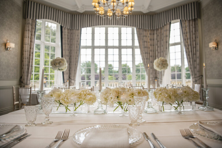 Crystal glasses and white flowers on a wedding table by a tall window, De Vere Horsley Estate