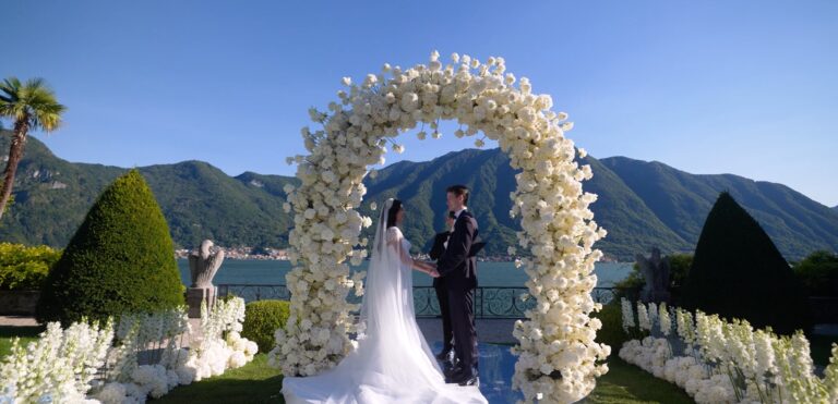 A couple under a white floral arch at Villa Balbiano, with the mountains of Lake Como behind, Denee Motion