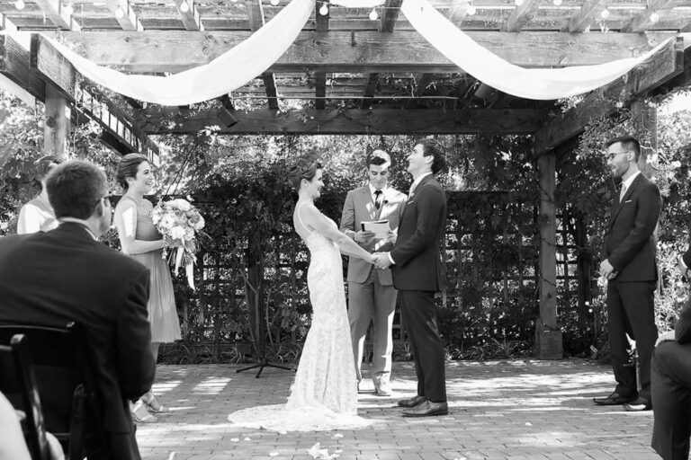 Vows beneath a pergola draped with white fabric, guests gathered around, in black and white, Despina Craig Events