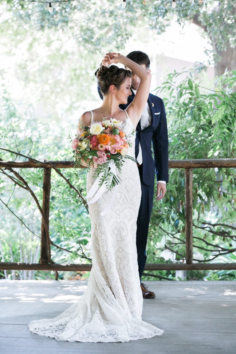 A bride in a lace gown holding a peach bouquet, with her groom, on a timber deck, Despina Craig Events