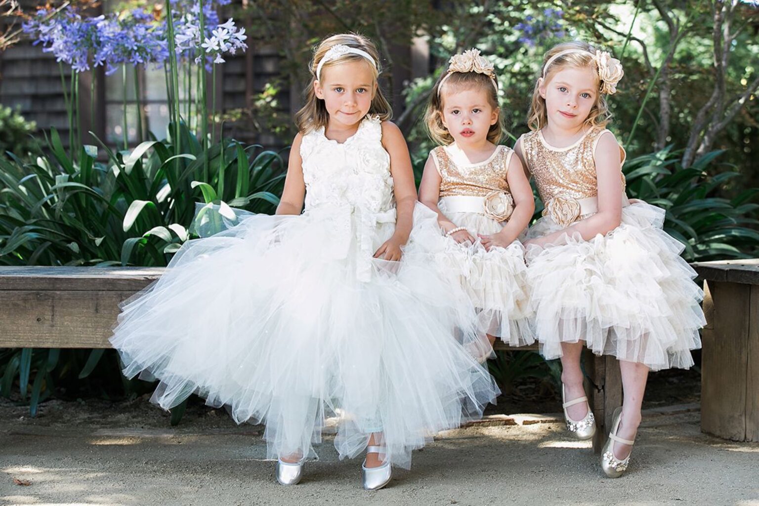 Three flower girls in tulle dresses seated on a garden bench, Despina Craig Events