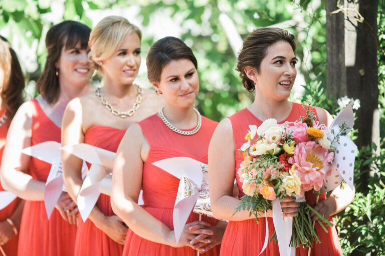 Bridesmaids in coral dresses holding pinwheels, with a bright bouquet in front, Despina Craig Events