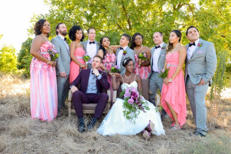 The full wedding party grouped in pink and grey on a dry-grass lawn, Despina Craig Events