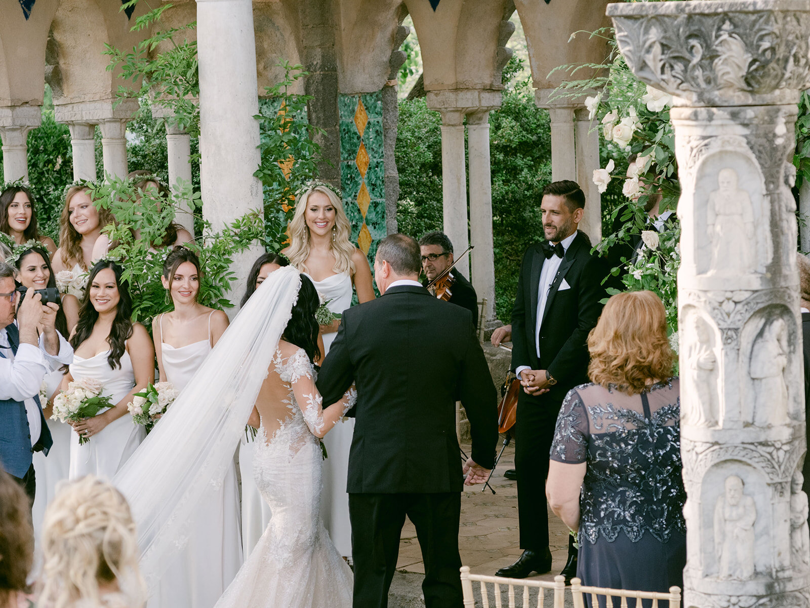 The bride arriving among guests and bridesmaids beneath the carved stone arches. Alex and Andrew's wedding at Villa Cimbrone, Ravello, Amalfi Coast, Detito Photography