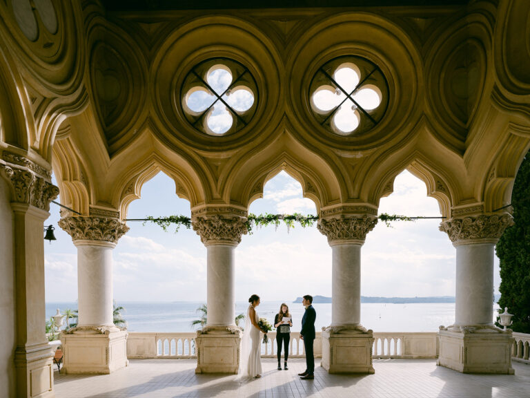 The ceremony framed by pointed stone arches with the lake beyond. Sam and Lance's elopement on Garda Island, Lake Garda, Detito Photography