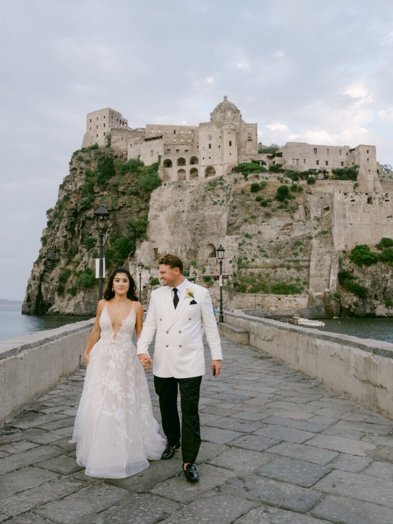 The couple walking the stone bridge with the castle rock rising behind. Erica and Dustin's wedding at the Aragonese Castle, Ischia, Detito Photography