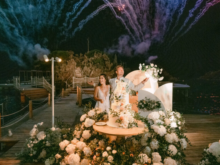 Cutting the cake beside a bank of white flowers as fireworks open over the water. Erica and Dustin's wedding at the Aragonese Castle, Ischia, Detito Photography